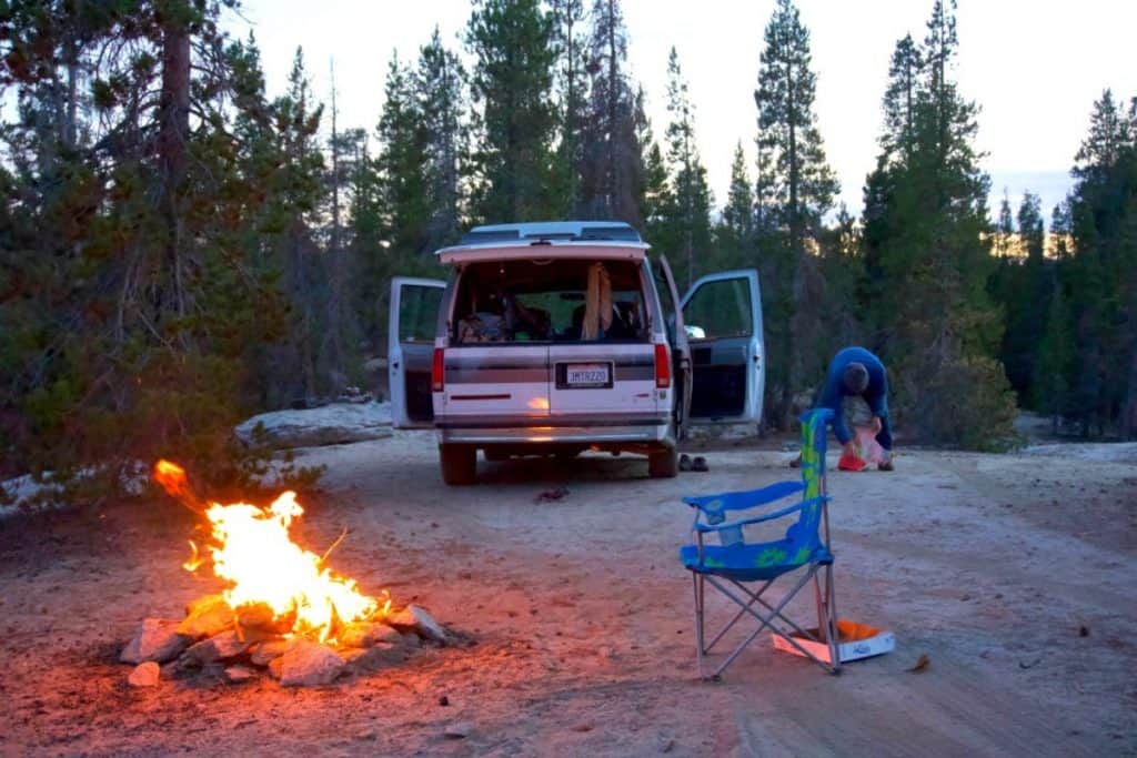 The van is camped out for the night, there is a fire bruning large with a chair in front of it. They appear to be camped in the middle of the woods.