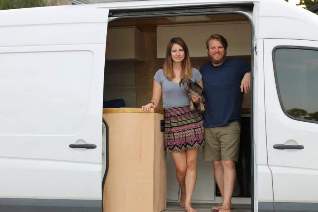 Girl and guy stand inside their Sprinter van built to be a tiny home. The side door is open and they stand in the opening next to their kitchen counter.