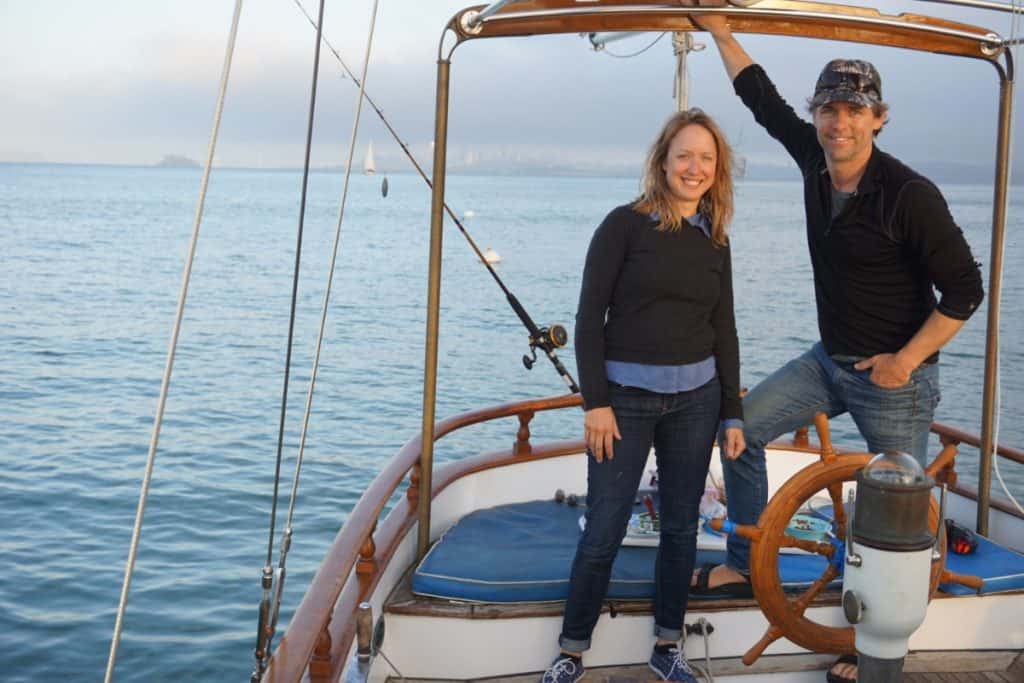 Girl and guy stand on their sailboat. There is a fishing pole behind them.