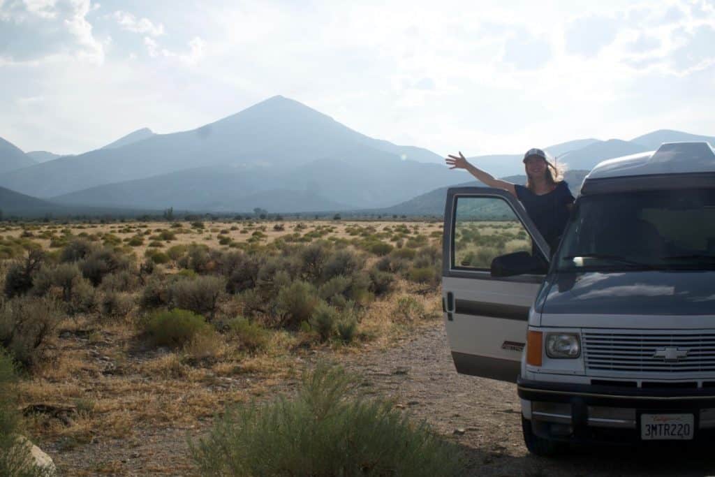 Girl waves outside of the passenger side of the van. There is a large mountain in the background.