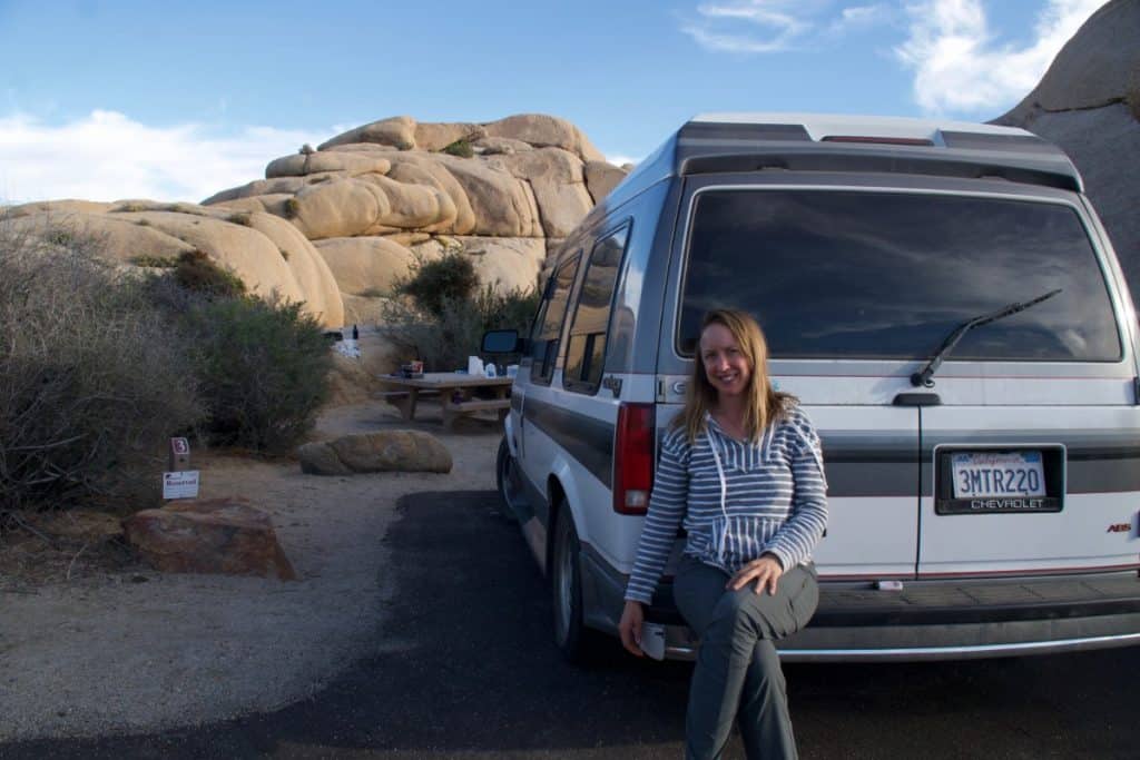 Girl sits on the bumper of her van with a large rock formation behind them.