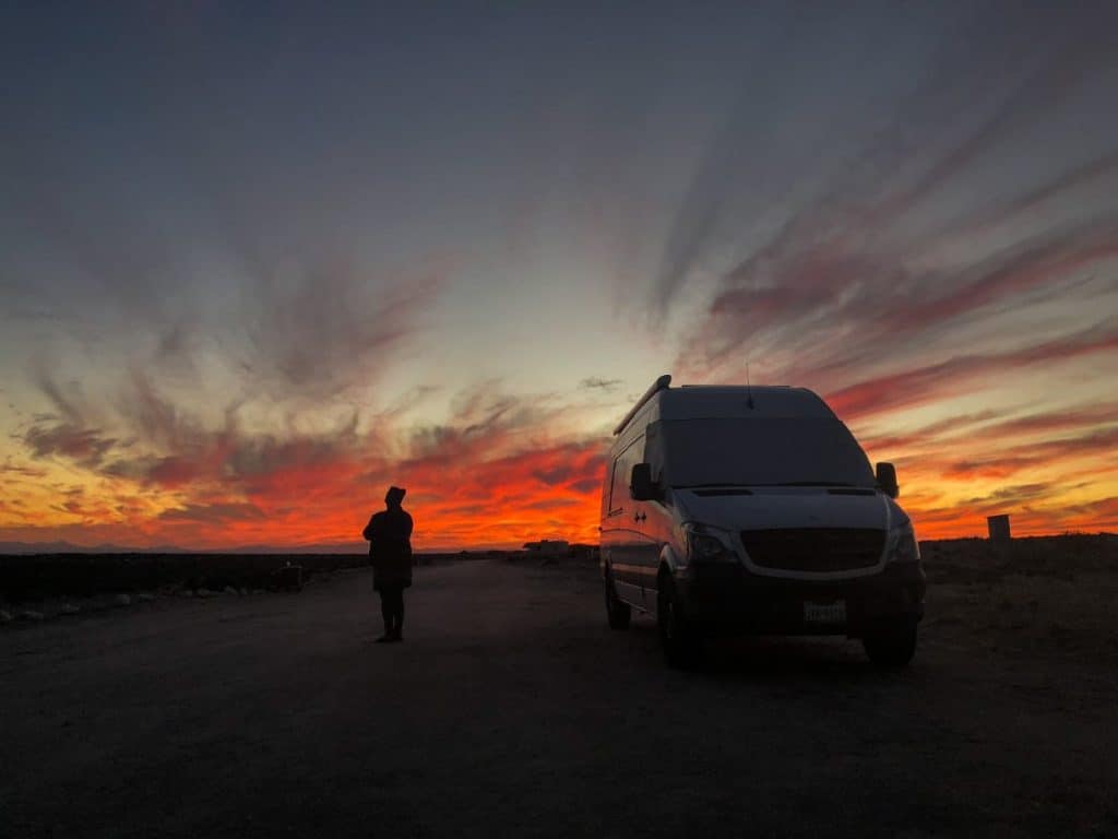 The sunsets behind the white Sprinter van with vibrant oranges and reds.