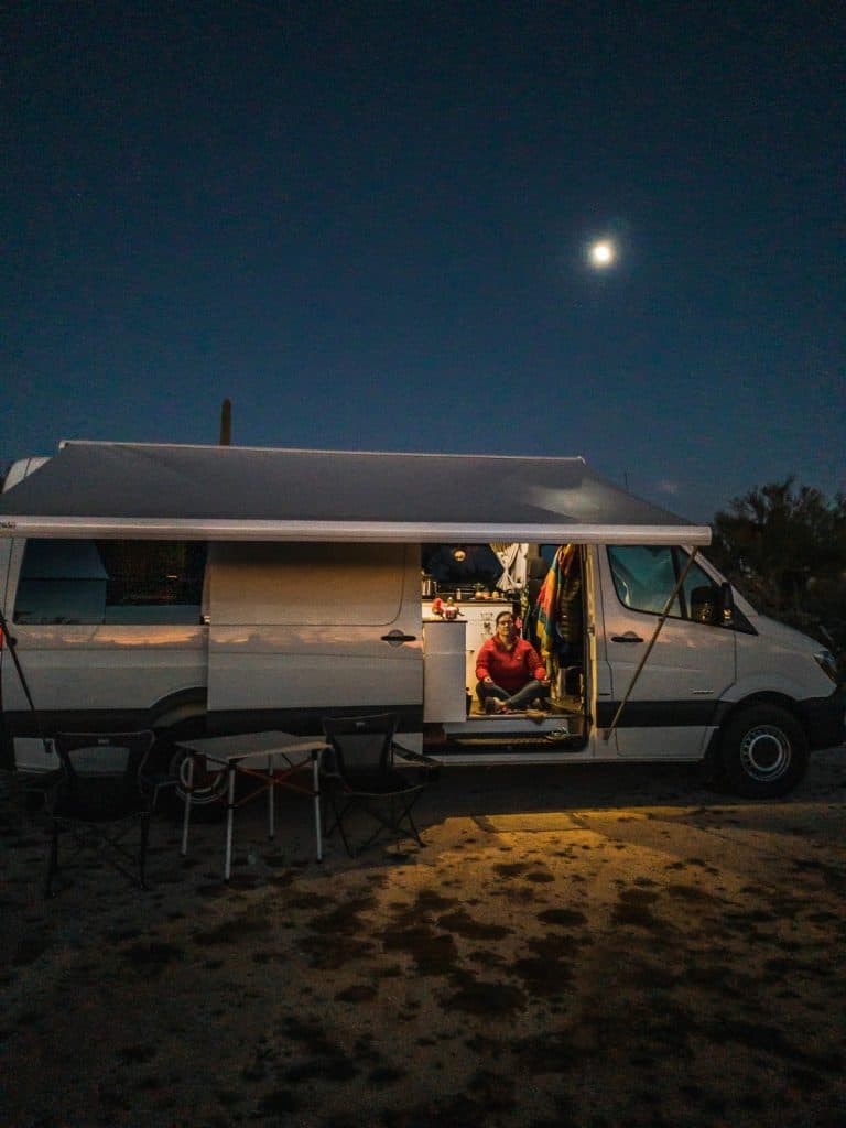 The van is parked out at night with it's awning stretched out and side door open. Meghan meditates in the open doorway as the moon shines bright high above the van.