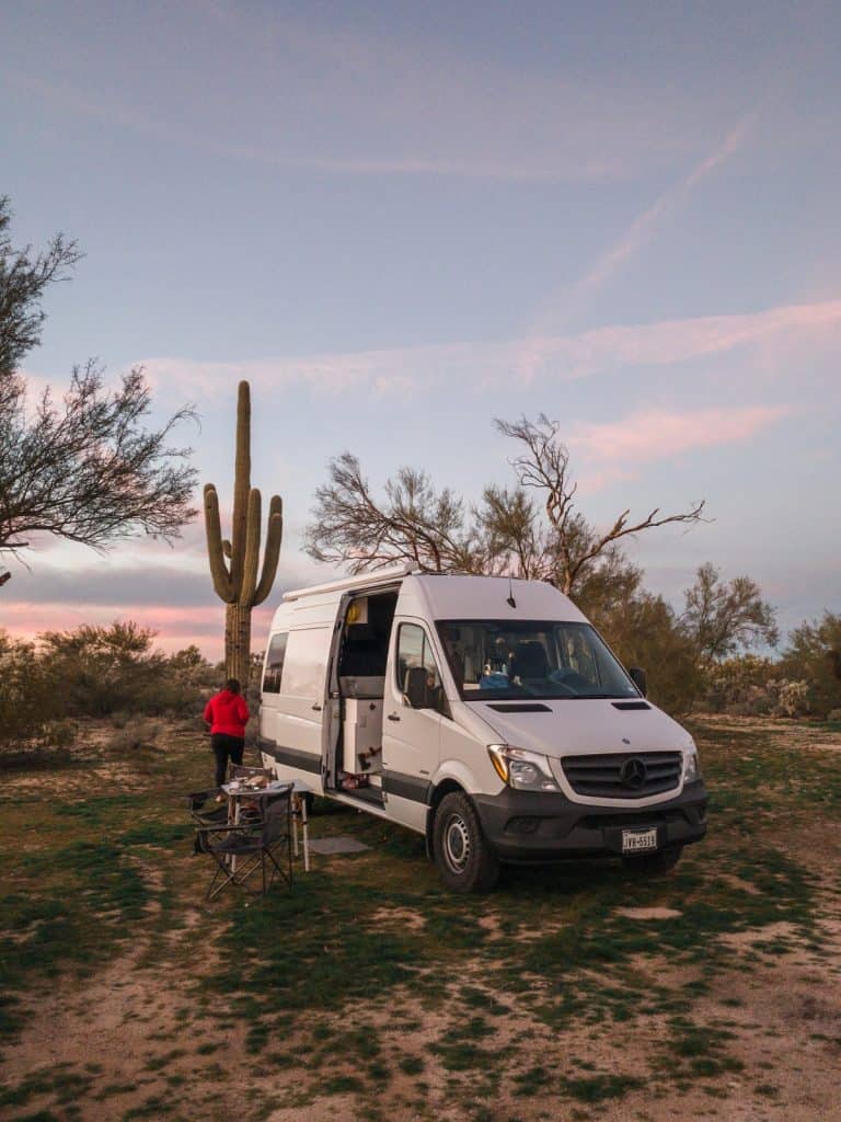 The van is camped out in the desert with a large saguaro cactus directly behind it. The side door is open, a table and two chairs sit out next to the side doors of the van.