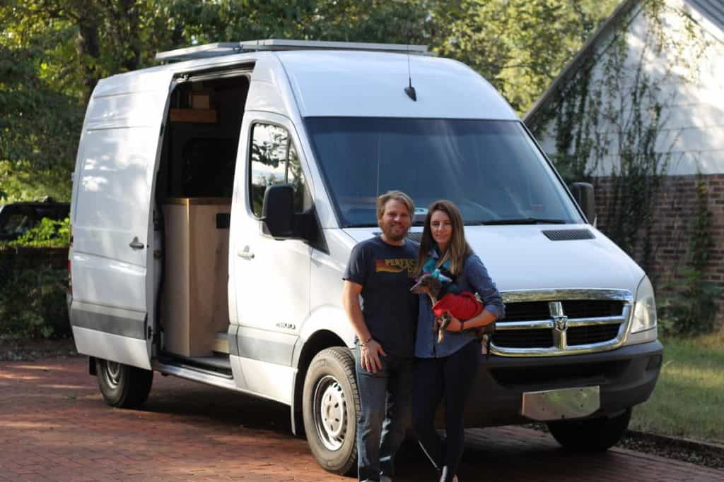 Guy and girl stand in front of their sprinter van holding their dachshund.