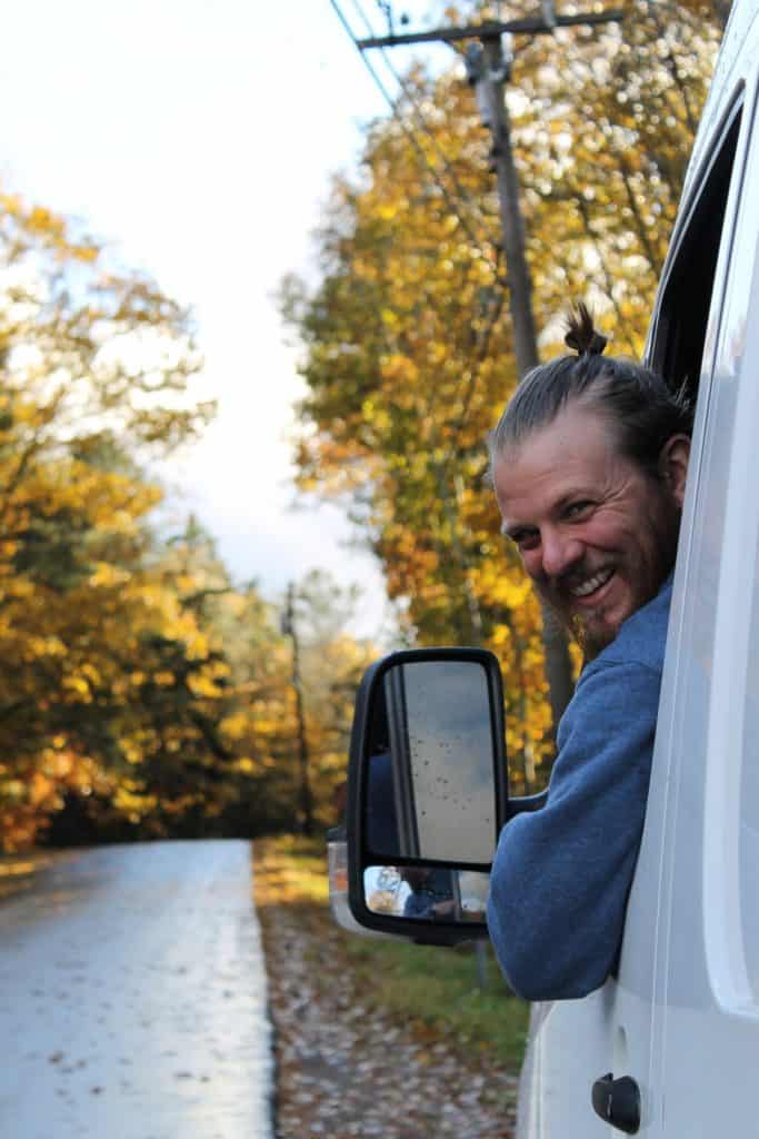 Guy leans out of driver side window looking back at the camera and smiling. Autumns changing leaves sparkle in the background.