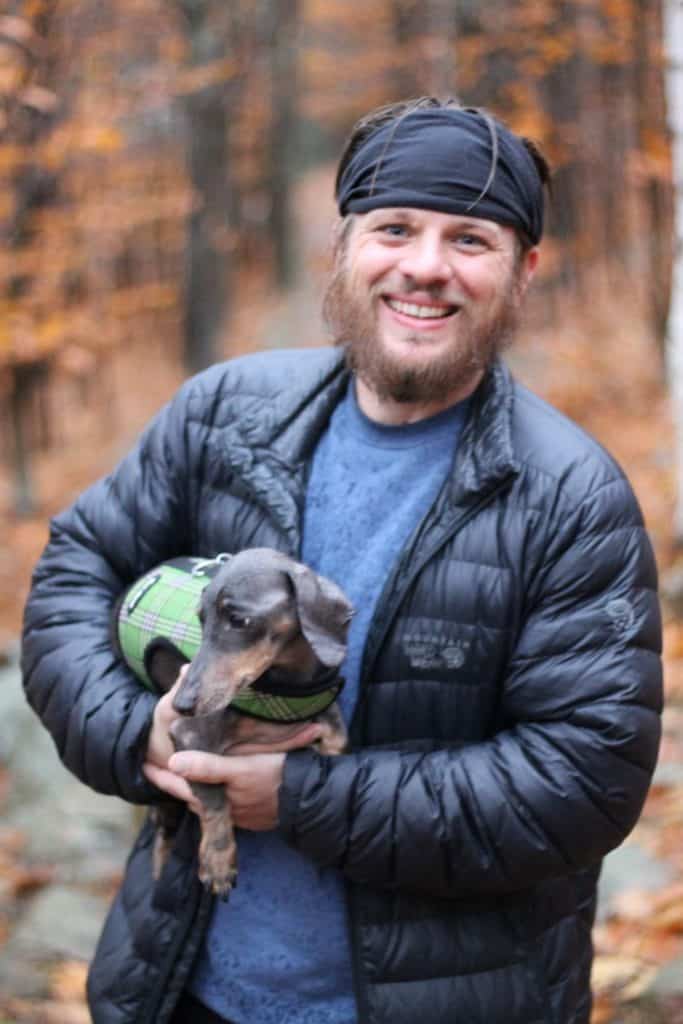 Guy stands in the woods holding his dachshund looking happy.