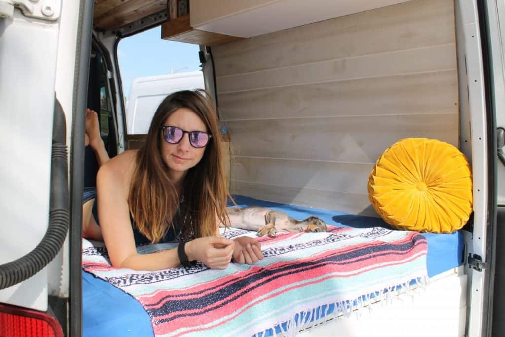 Girl lays in bed with the back doors of her van open. She wears sunglasses as the sun beams down on her and her dachshund passed out in the bed next to her sunbathing as well.