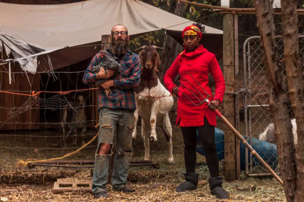 Man, woma, and goat stand facing the camera. The man holds a chicken and the woman holds a pitchfork. The goat looks condescendingly forward.