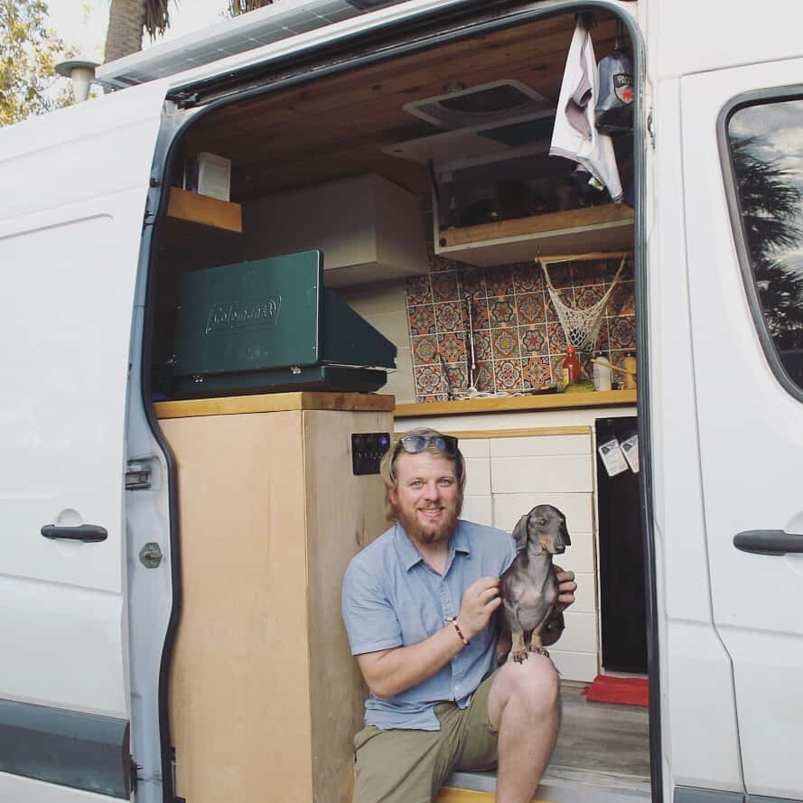 Guy sits on the side entrance of the van with his dachshund on his lap.