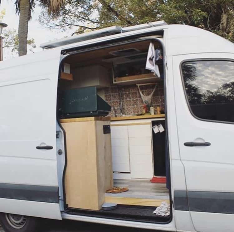 The side door to the Sprinter is open and we are able to see inside their kitchen. We see a stove propped up, cabinets, a fruit hanging basket, and little knick knacks.