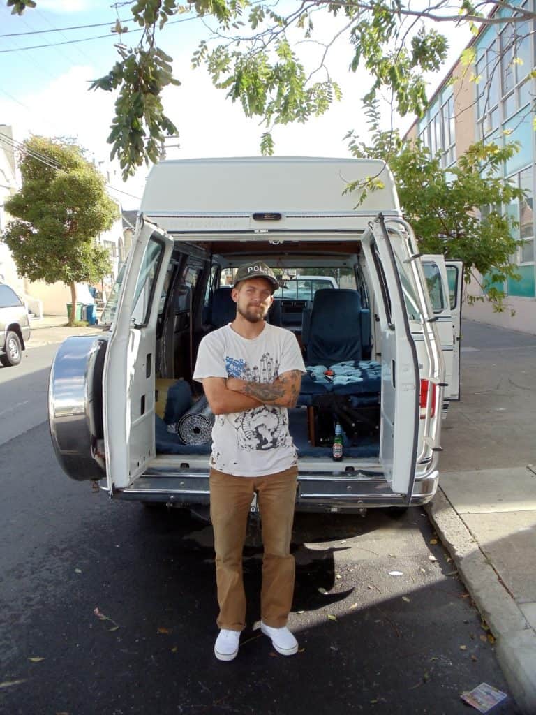 Guy stands in front of van, the van seems to not be built quite yet but has materials inside.