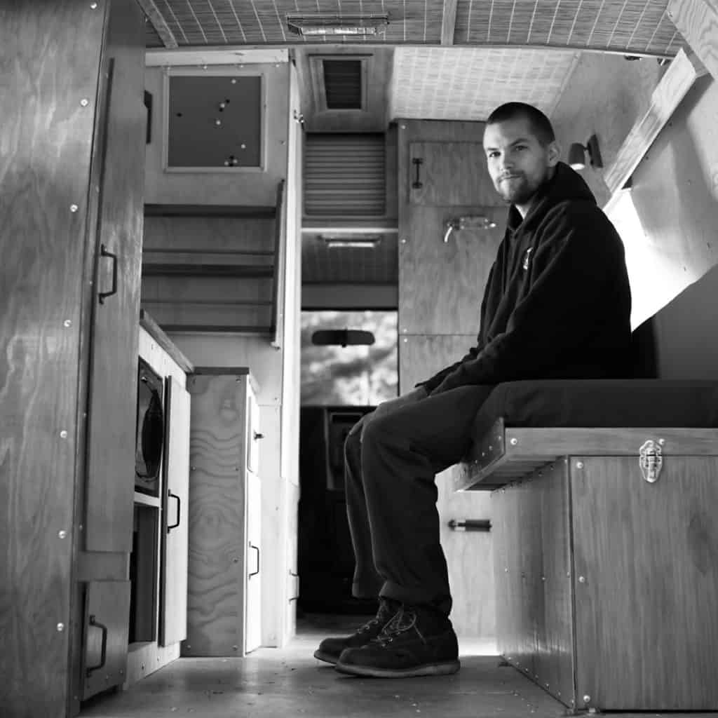 Black and white shot of a guy sitting on a couch inside of a van built out to be a tiny home.