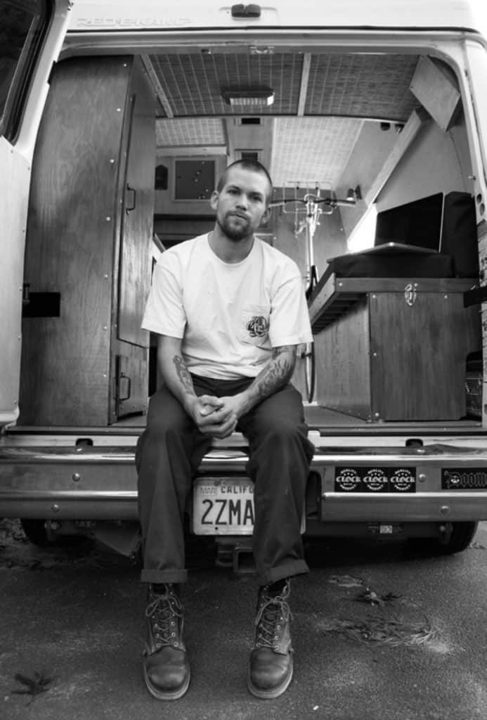 A white man with tattoos sits in the back of a van he built to be a tiny home. The image is black and white.