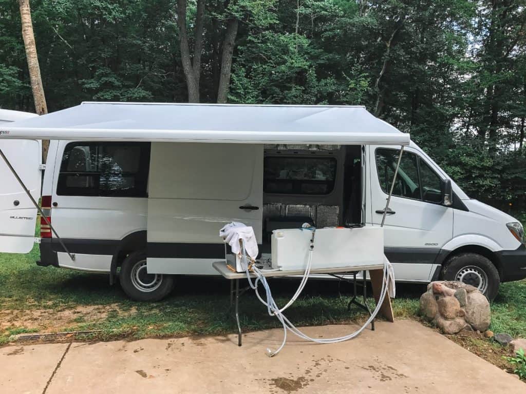 A side photo of the van. A table sits outside the van holding their water tank, hoses and additional plumbing equipment.