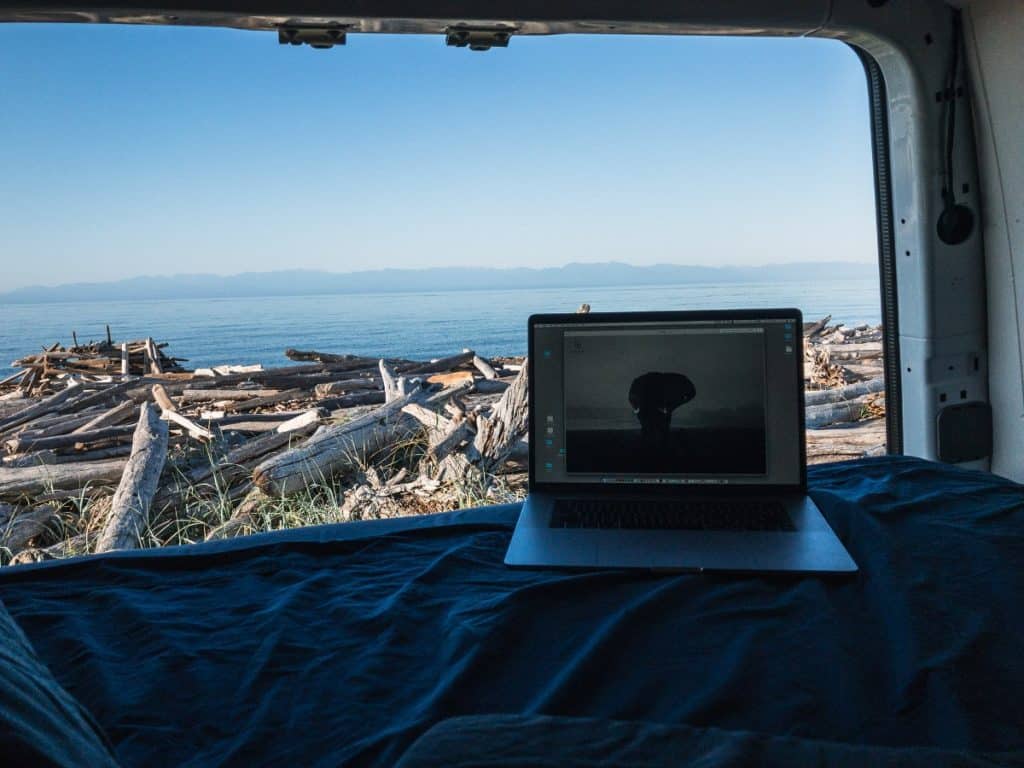 A TV sits on the bed. The back doors are open and we see a view of driftwood and a vast amount of water, probably an ocean.