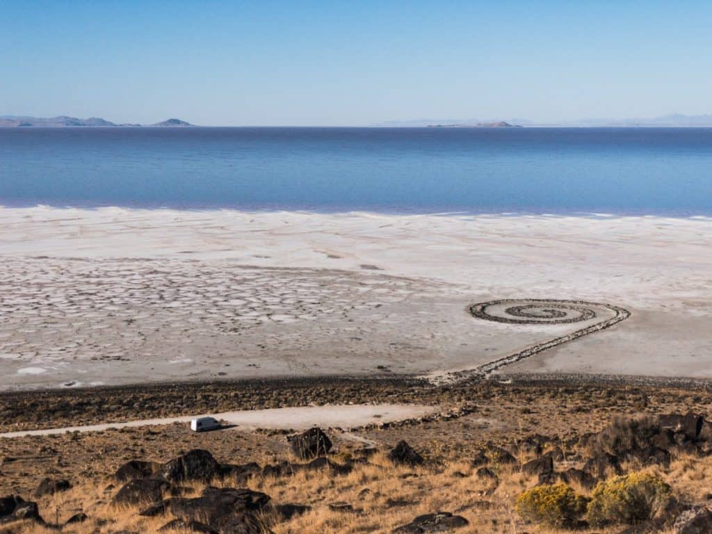 This photo is taken up a mountain looking down. Far off in the distance we see the van. Just past the van we see a zen rock spiral trail. Just past the trail appears to be an ocean.