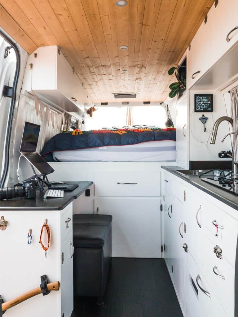 An interior shot of the van. We see cabinets off to the right where the kitchen area is with an induction stove, sink and faucet. Straight ahead is a raised queen sized bed with storage underneath. To the left is a desk with drawers and a propped up laptop.  A perfect little workspace.