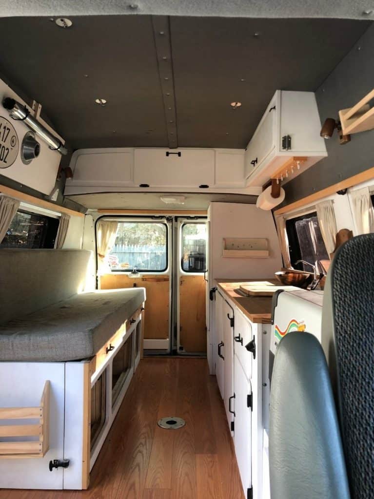 Interior shot of a van built to be a tiny home/ There is a couch to the left, a long walkway splits up the space, and the right is the kitchen counter. The top and bottom are filled with different types of cabinet storage.