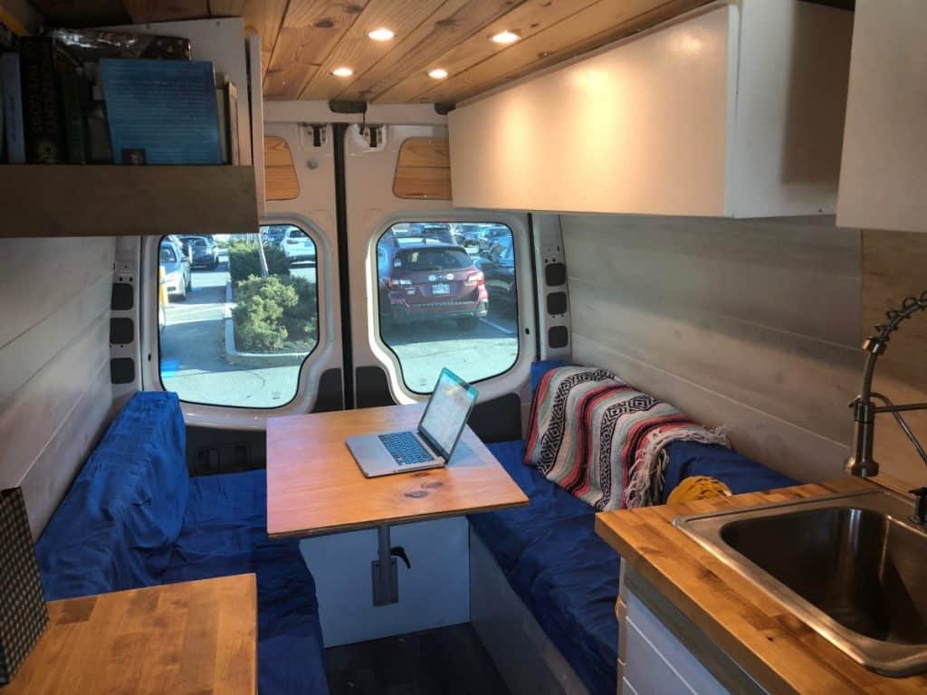 A view of the inside of the van near the rear. There is a couch on each side of the van with a table top in the middle. Two cabinets sit up near the ceiling on each side of the van.