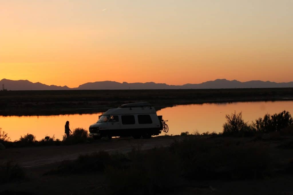 Van is parked on a beach near water as the sun sets over mountains in the distance.