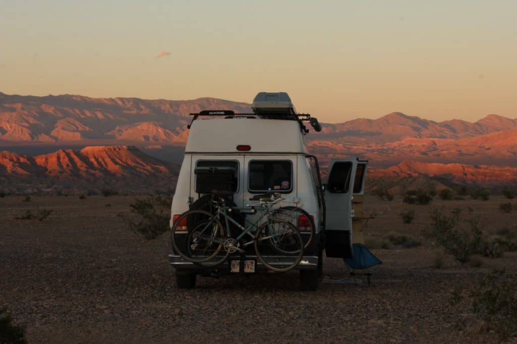 A shot of the back of a van. There are bikes mounted to the back. The side doors of the van are open and there is a chair on the ground in front of the doors. In the background of the shot is the sun setting over the beautiful red rocks of Nevada's mountains.