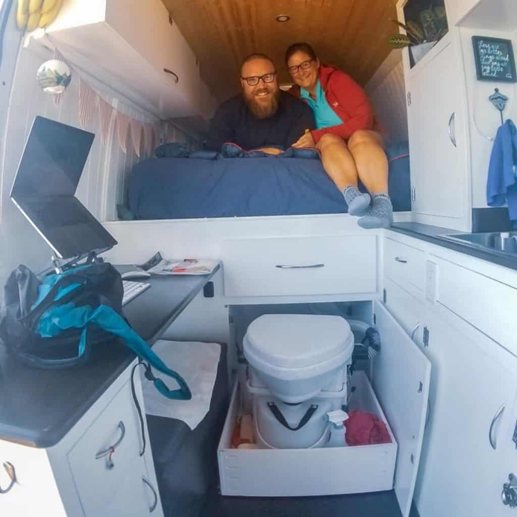 The happy couple poses on their queen sized bed. Below the bed, a storage door is open and their Nature's Head composting toilet slides out to expose how it can come out when need be, or slide back into hiding when not being used.