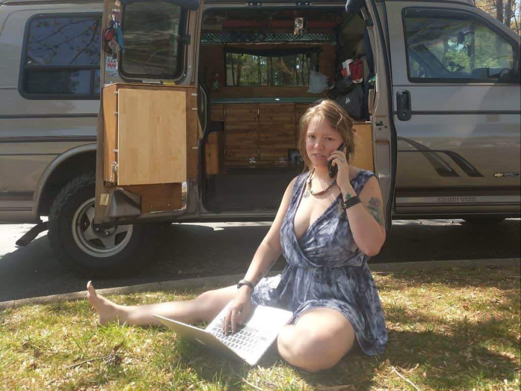 Jayme sitting in the grass in front of the van, on the phone, typing at a computer, looking busy.