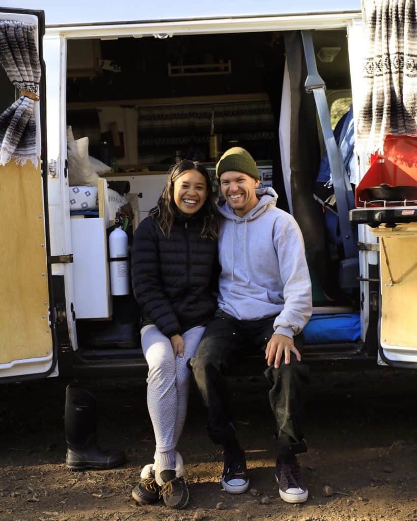 Girl and guy sit on the edge of their van where the side doors open. The van is built to be a tiny home. They both grin wide and happily.