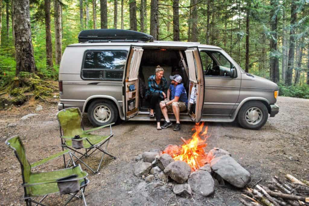 A girl and guy sit on the edge of their van where the side doors open. They are in the middle of the woods and a large campfire roars in front of them.