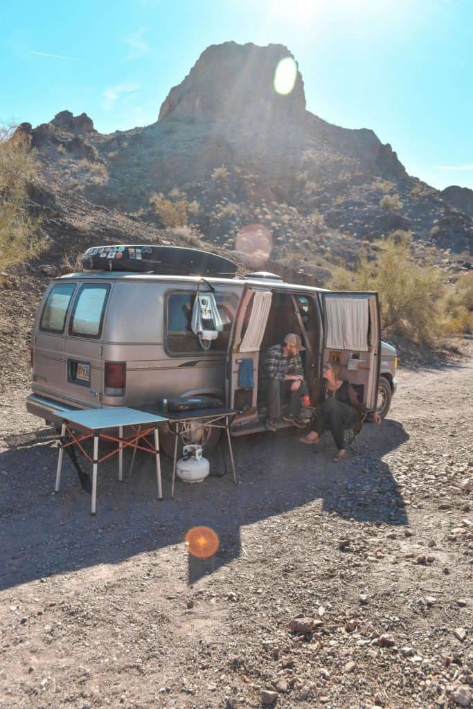 Guy and girl sit outside of their van with the side doors open and a large mountain view behind them. They have tables and a stove sitting out as well.