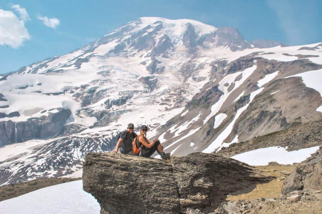 Guy and girl sit on a rock ledge with the peak of a snow capped mountain directly behind them.