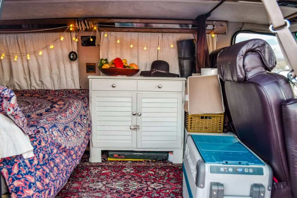 The interior of a van built to be a tiny home. It is a shot from the side doors looking in. We see a fridge behind the passenger seat and a dresser and some additional storage behind the drive seat. The bed begins on the left.