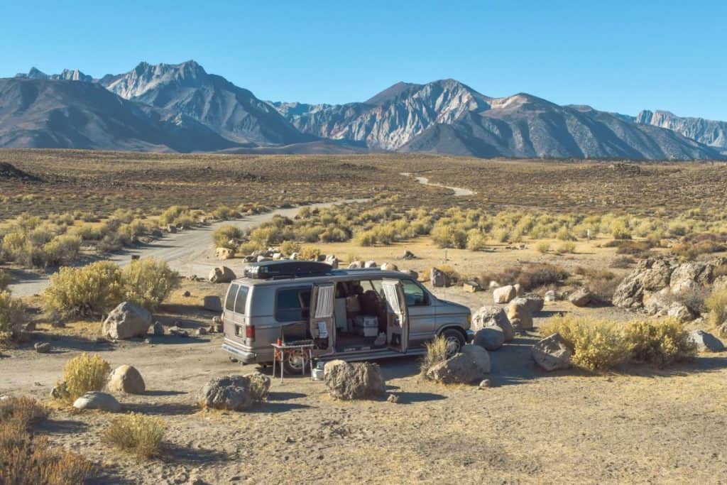 A van camps out in the middle of a desert with epic mountains behind it. The van is camped out with a table out front and the side doors open.