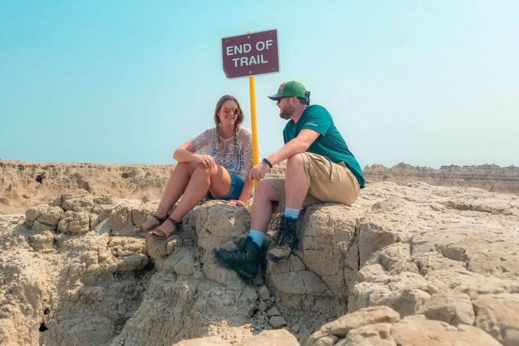 Girl and guy sit on small rock formation next to a sign that says "END OF TRAIL".