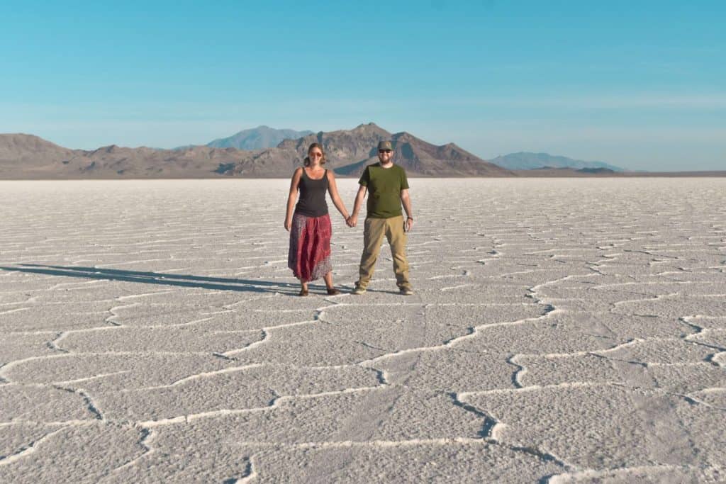 Girl and guy hold hands standing on salt flats with mountains behind them.
