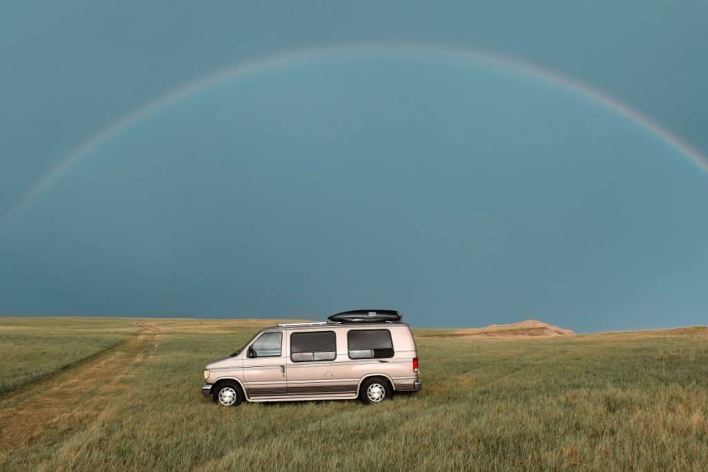 A van sits in the middle of a field with a rainbow arching directly over it.