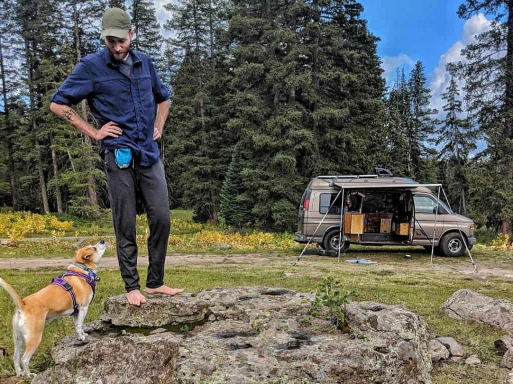 man stands on rock with dogs sniffing below him and van in the background