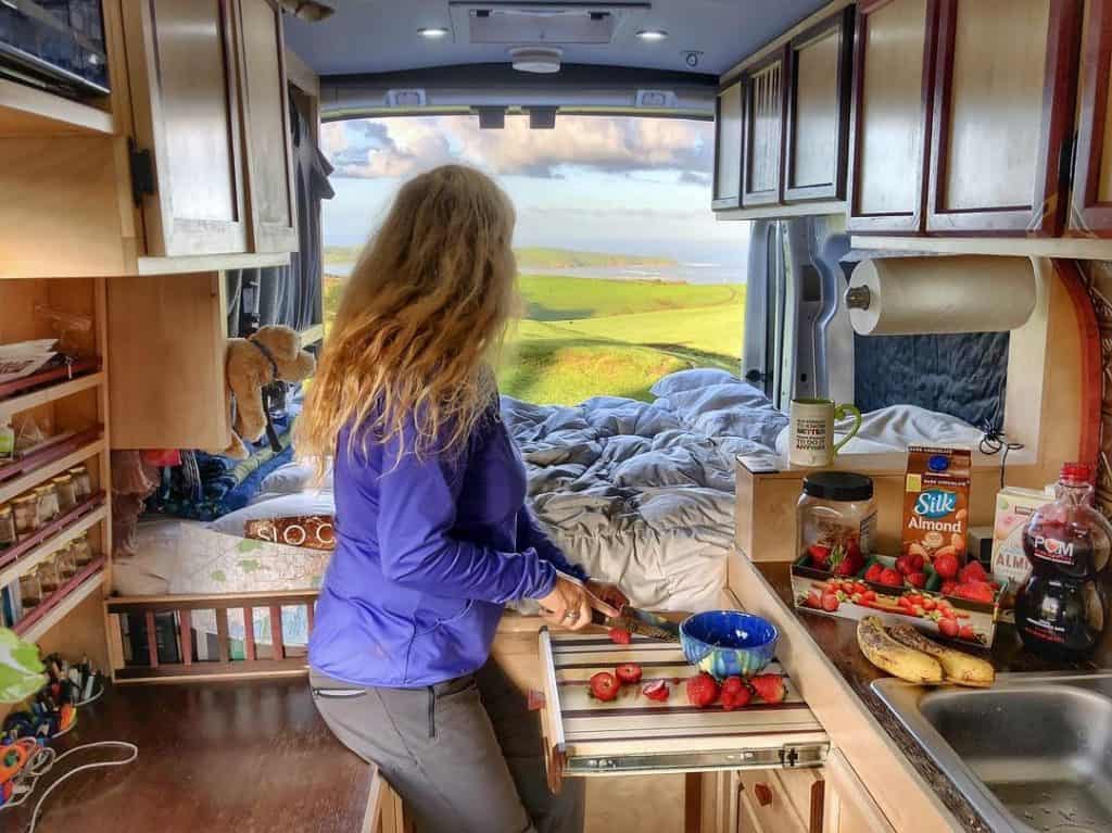 woman cooking in her van looks out the backdoors to a beautiful landscape