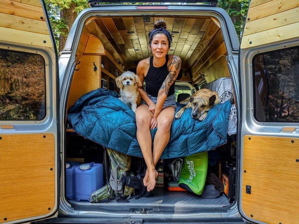 woman sits on the edge of a bed with her dogs in her van