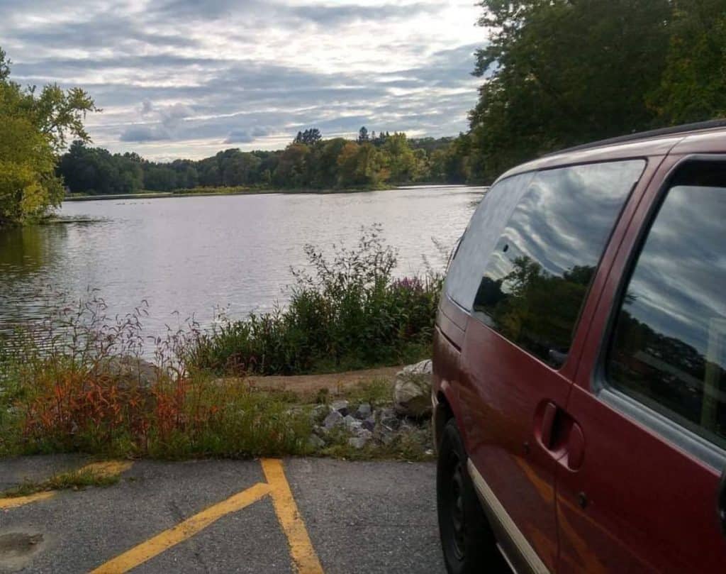 A red minivan is parked with a lake behind it
