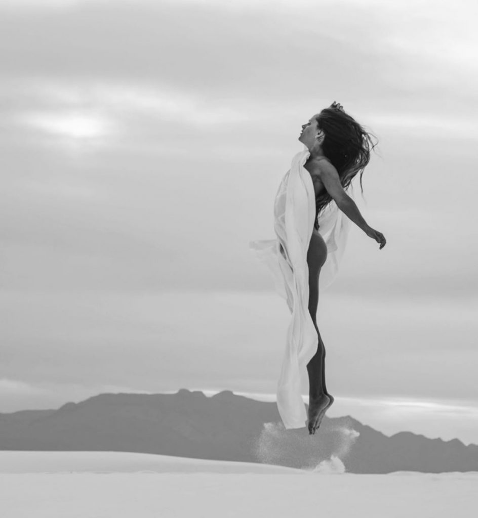 A black and white photo of a woman jumping up in the air (almost looking like she's floating) wrapped in a white sheet.