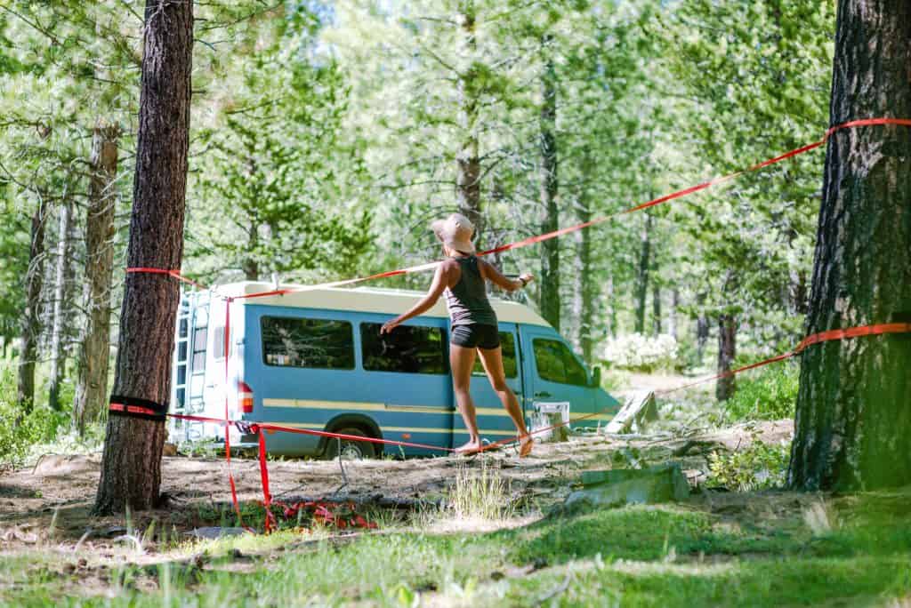 Nat walks across a slackline in the woods with the van in the background.