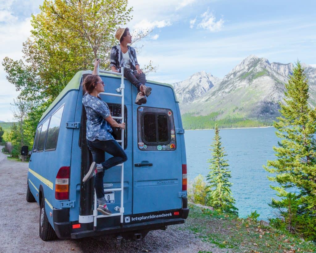 Nat sits on the roof while Abi hangs out on the ladder on the back. They are looking over a lake and mountains.