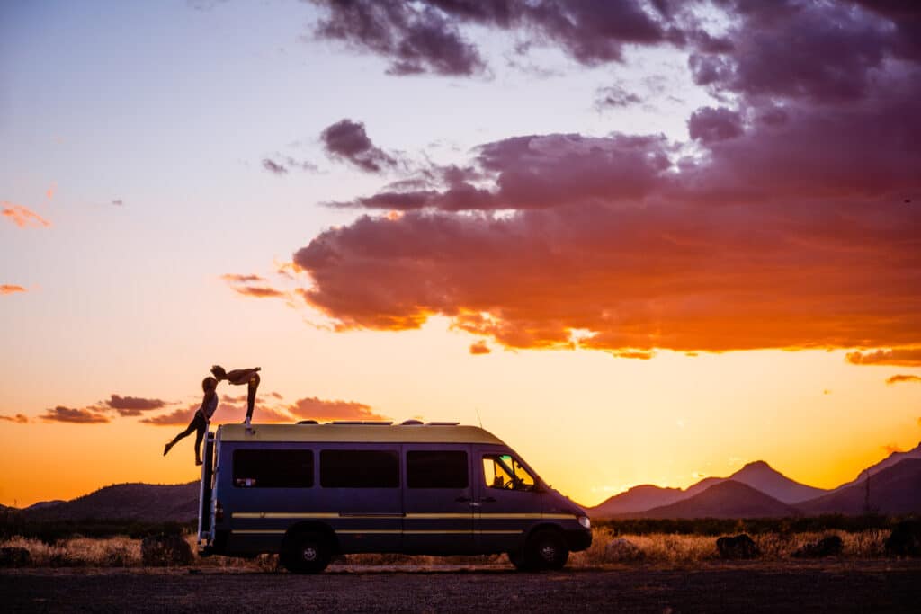 Nat stands on top of the van, bending over to kiss Abi who is climbing her way up the ladder. The sun is setting and there are Southwest mountains around them.