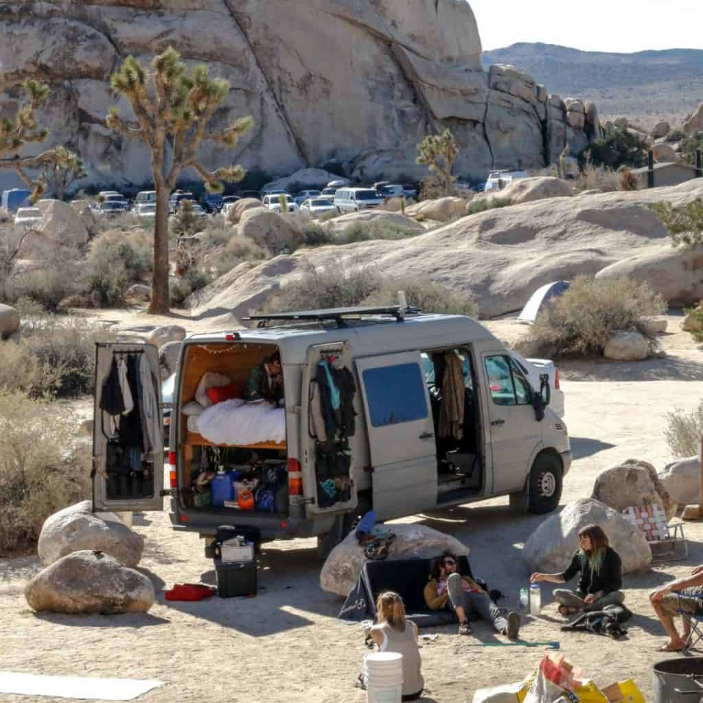 A van is parked surrounded by rocks and boulders with people hanging out outside of the van.