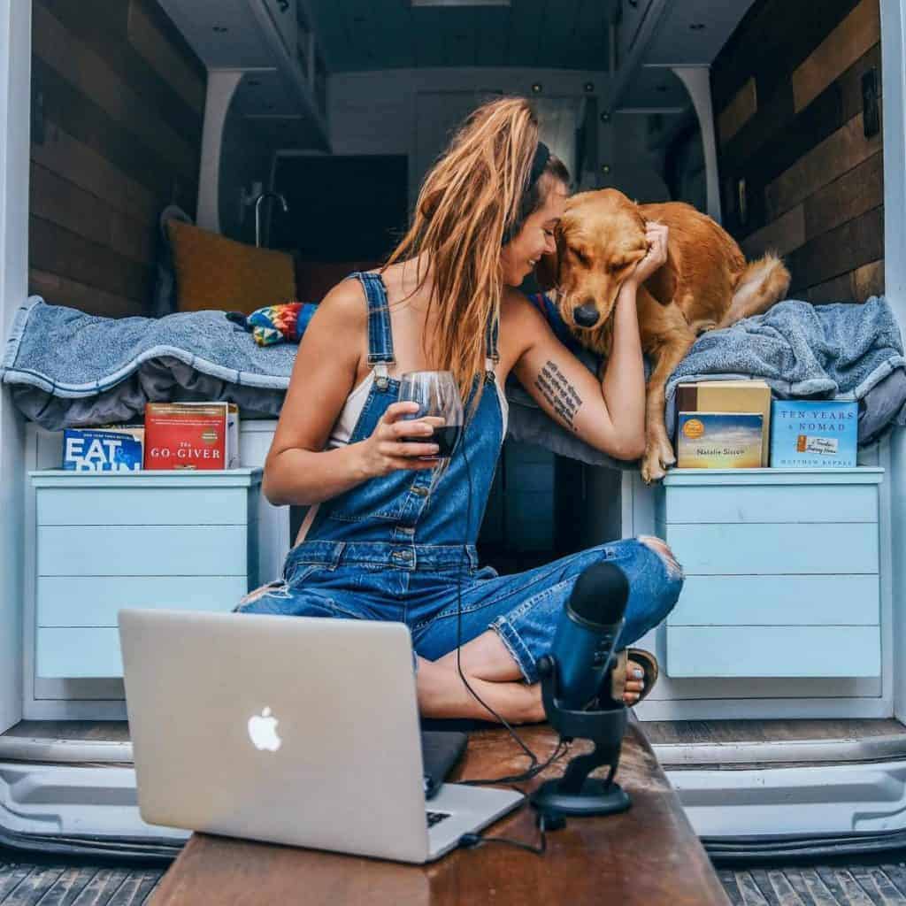 Sydney sits on her table that pulls out the back, petting her dog, holding a glass of wine, and working on her podcast.