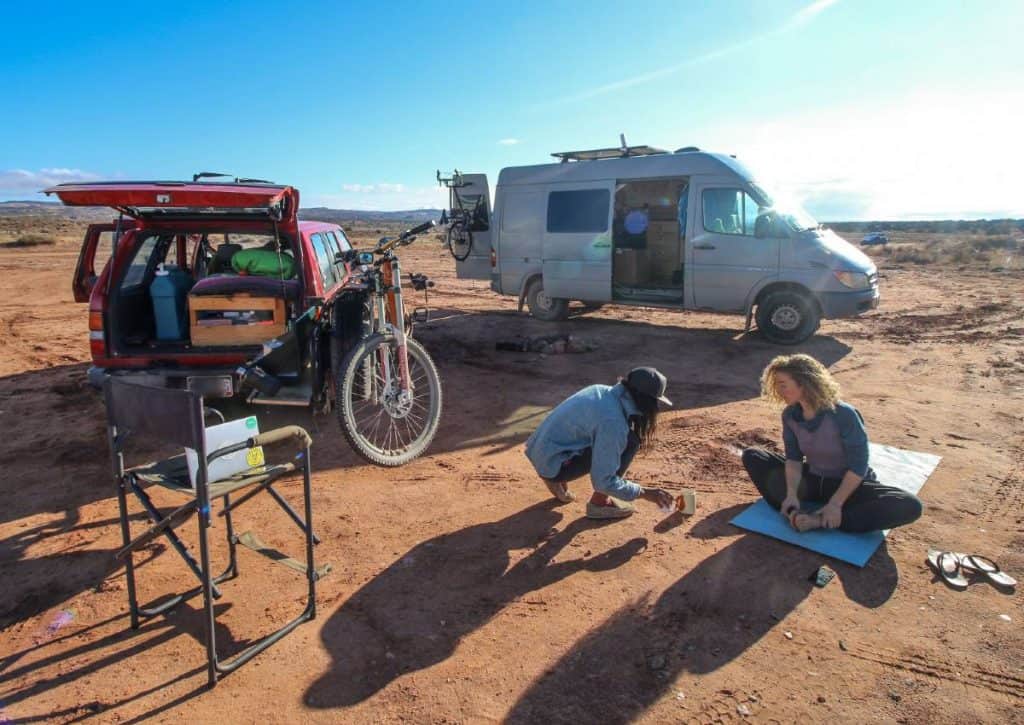 A van and an SUV are parked on flat dirt land. Two people hang out outside on the ground.