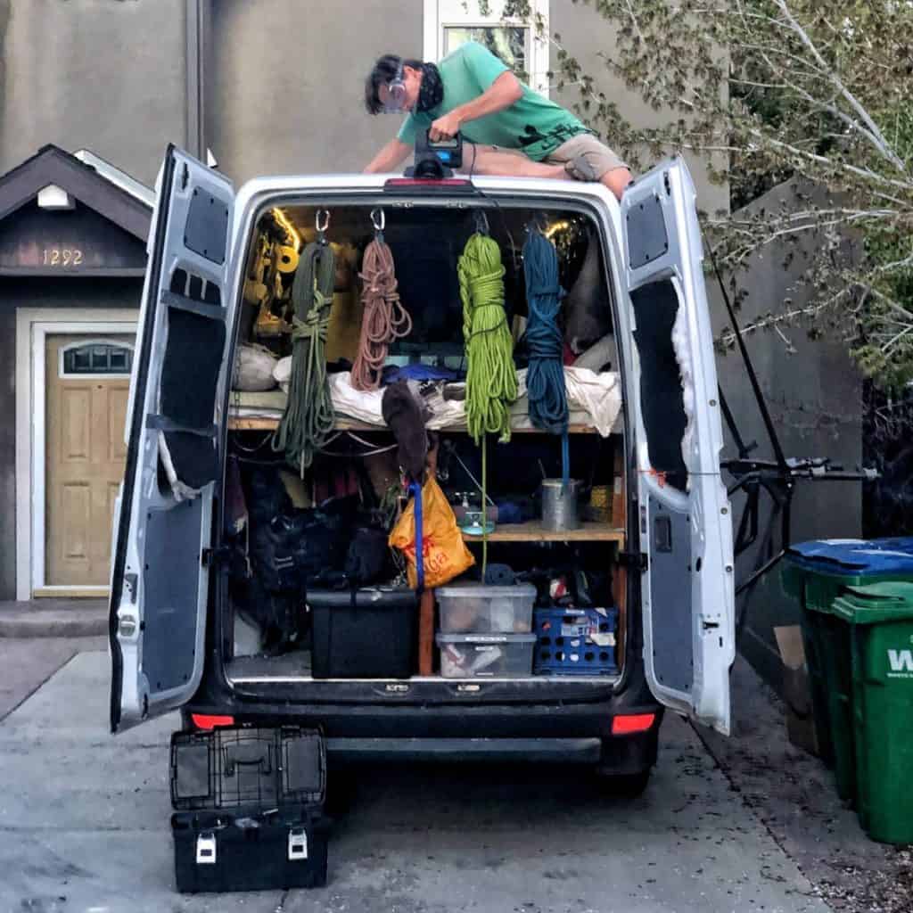 Someone is working on the roof of a van with the van's back doors are open. The back doors expose ropes and other rock climbing gear.