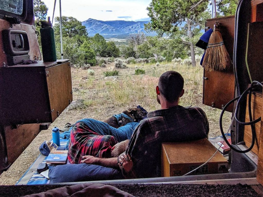 Jayme John and Nymeria cuddle outside of the van's side doors, under their awning, looking at a mountain range.